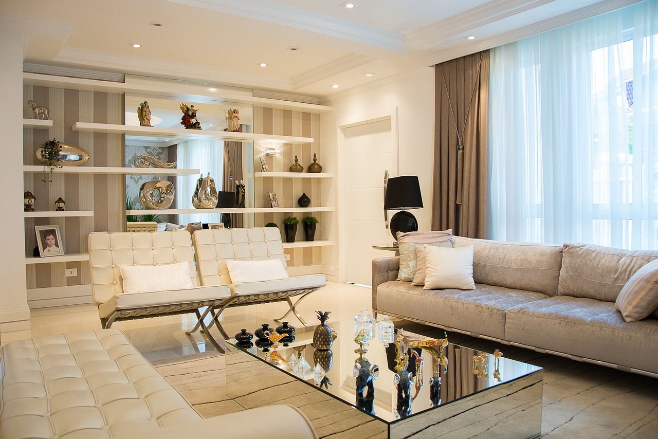 ห้องนั่งเล่นโมเดิร์นที่ตกแต่งด้วยโซฟาสีเทาขนาดใหญ่, เก้าอี้หนังสีขาว, ตู้วางทีวี, และชั้นวางของที่มีของประดับตกแต่งมากมายในโทนสีครีมที่สงบ